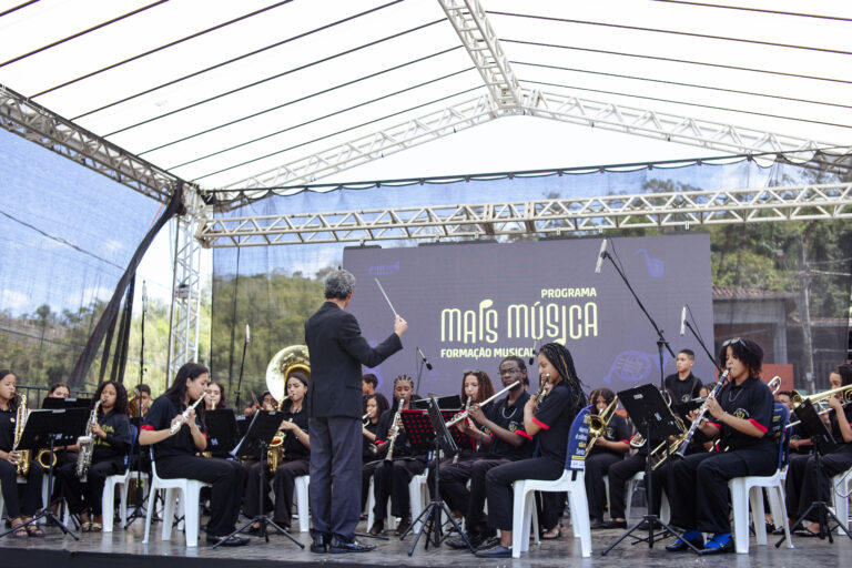Encontro de bandas de música durante evento em Antônio Dias, com maestro conduzindo orquestra ao ar.