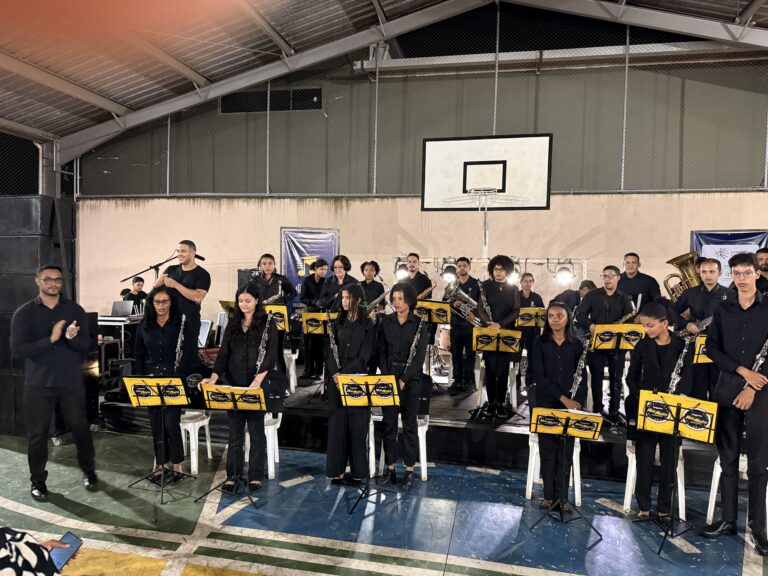 Encontro de bandas de música no Naque, com músicos tocando instrumentos em ginásio coberto.