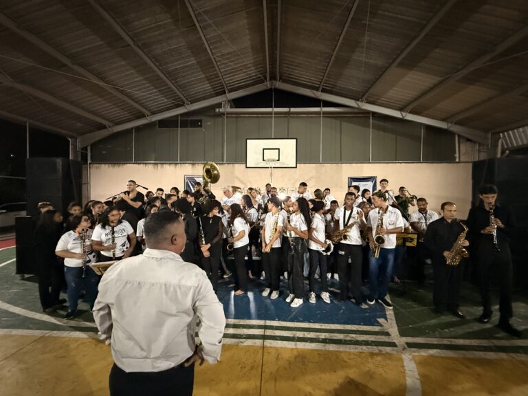 Encontro de bandas de música no ginásio de esportes de Naque, com músicos e instrumentos em destaque.