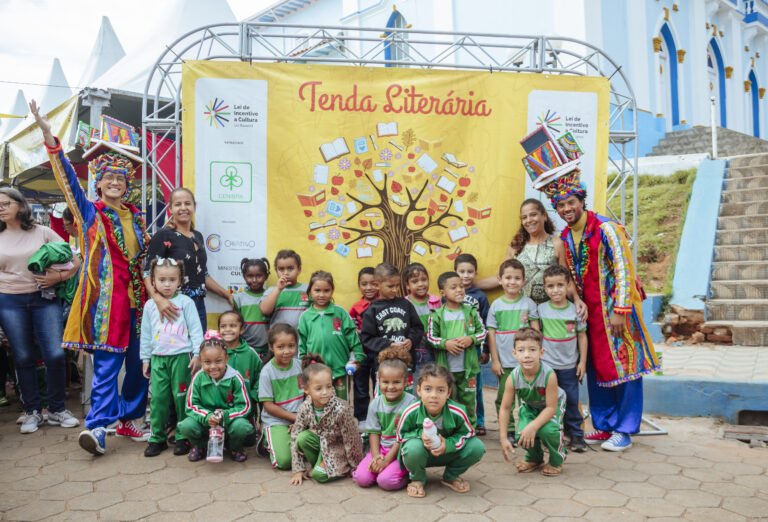 Imagem de crianças e adultos em evento literário ao ar livre com palco e decoração colorida.