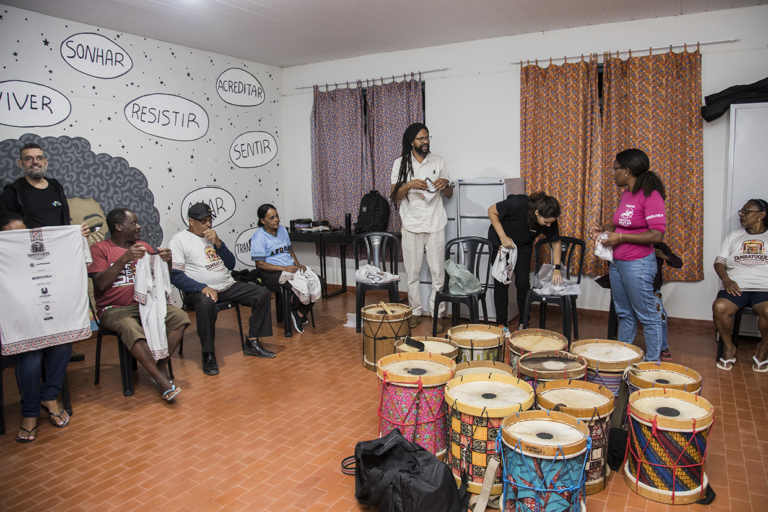 Aula de tambatuques com participantes aprendendo técnicas tradicionais em ambiente acolhedor.