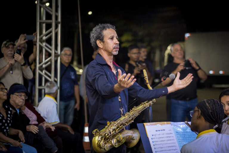 Músico conduz apresentação com saxofone em evento ao ar livre.