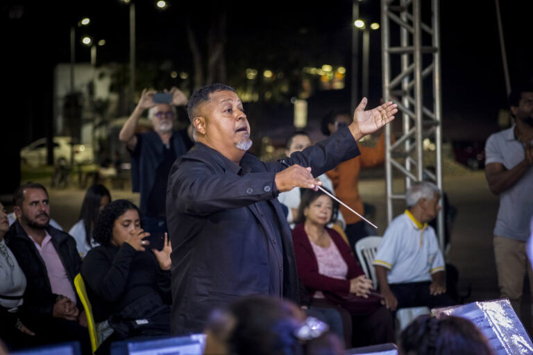 Homem conduzindo apresentação musical ao ar livre com público atento.