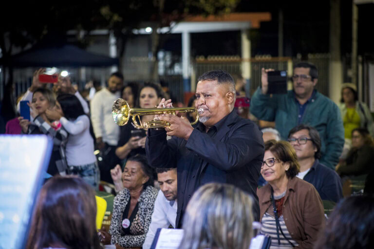 Músico tocando trompete em evento ao ar livre com público atento.