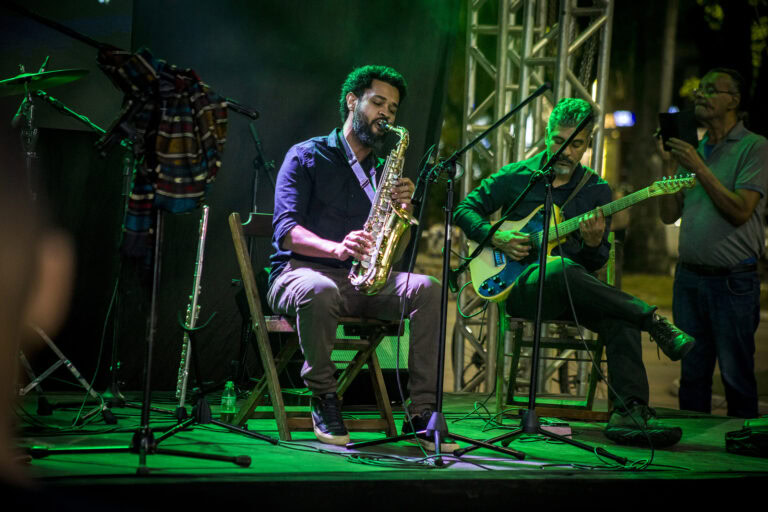 Músico tocando saxofone e outro na guitarra em palco ao ar livre.
