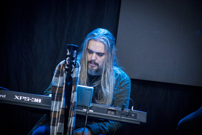 Músico com cabelo longo tocando teclado em show ao vivo.