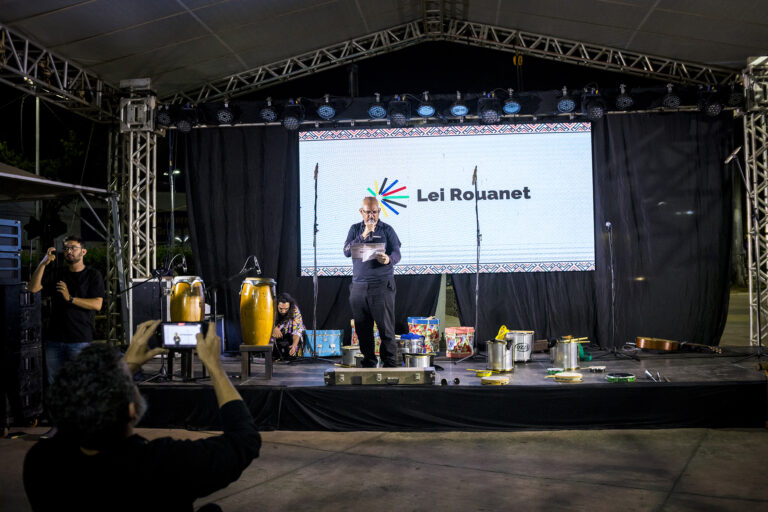 Homem falando ao microfone em palco no Festival Tambatuques.