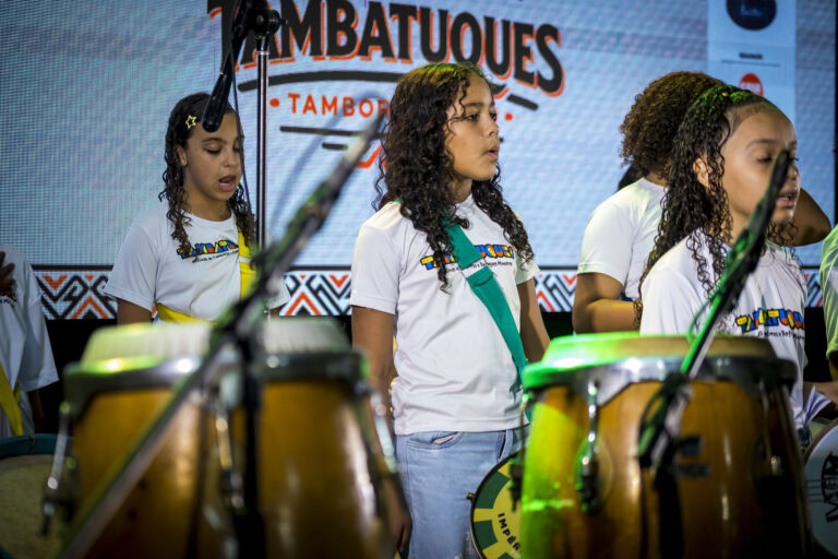 Crianças tocando tambor em festival de tambor com palco ao fundo.
