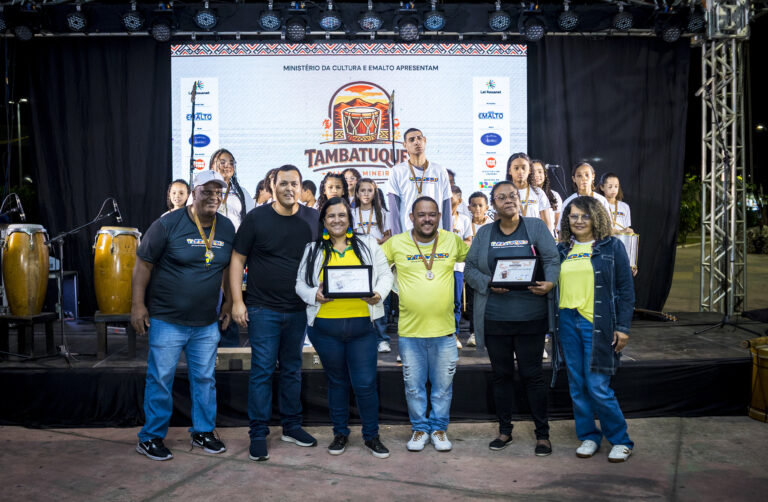 Grupo de participantes no Festival Tambatuques com palco ao fundo.