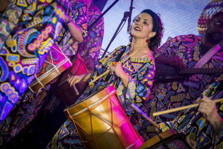Mulher tocando tambor em festival com roupas coloridas e vibrantes.